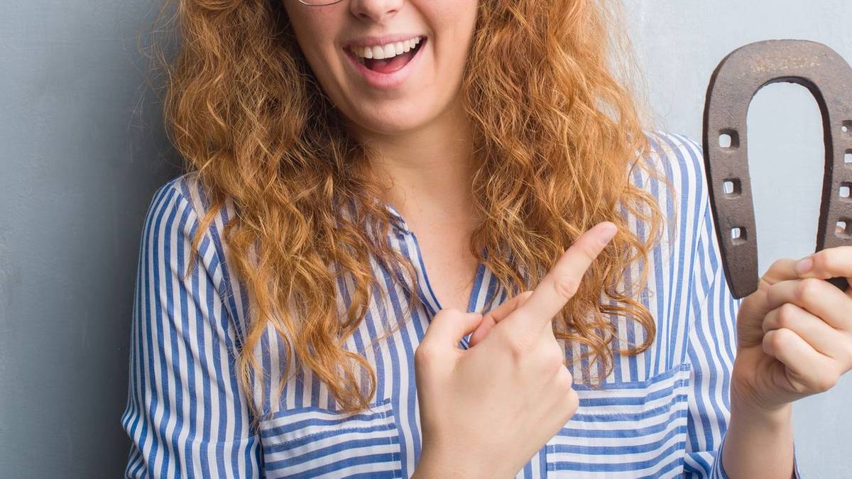 Uśmiechnięta kobieta z rudymi włosami i okularami wskazuje palcem na podkowę trzymaną w drugiej dłoni.