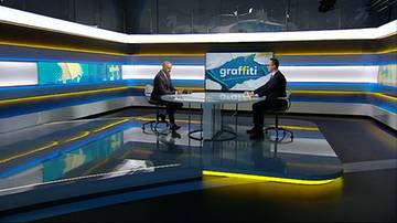 Dw&oacute;ch mężczyzn w garniturach siedzi przy okrągłym stole w studiu telewizyjnym.