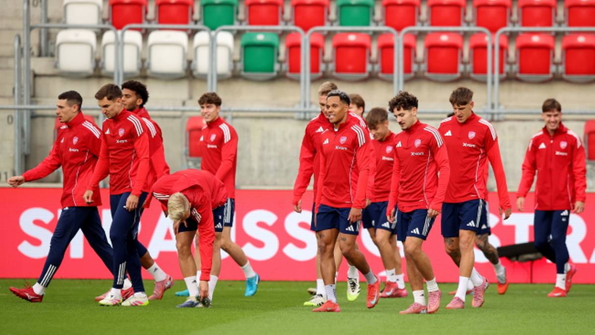 Grupa piłkarzy w czerwonych strojach treningowych idzie po zielonej murawie stadionu.