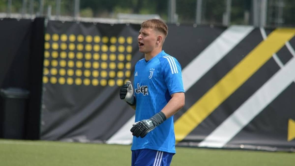 Młody mężczyzna w niebieskiej koszulce bramkarskiej Juventusu, z rękawicami bramkarskimi na rękach, stoi na boisku.