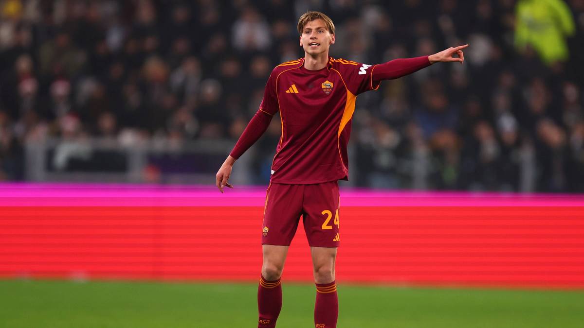 Piłkarz w bordowej koszulce z ż&oacute;łtymi detalami, numerem 24 na spodenkach, stojący na boisku i wskazujący palcem w bok.