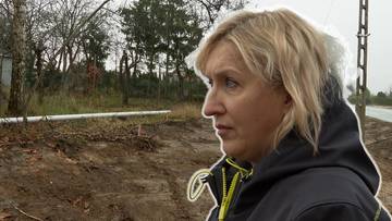 Kobieta o blond włosach patrzy w bok z zamyślonym wyrazem twarzy, na tle wyciętych drzew i terenu budowy.