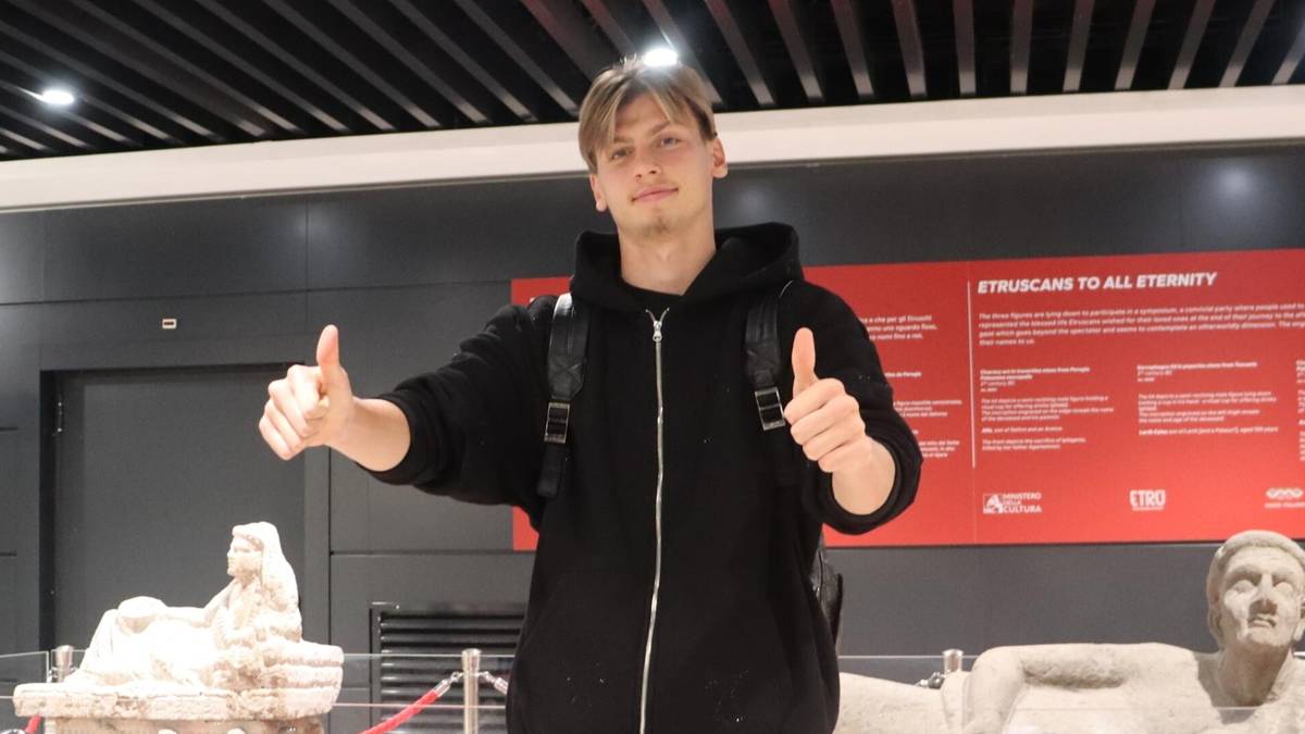 Młody mężczyzna w czarnej bluzie z kapturem i plecakiem na ramionach, wykonuje gest kciuka w górę obiema rękami. Uśmiecha się do aparatu.