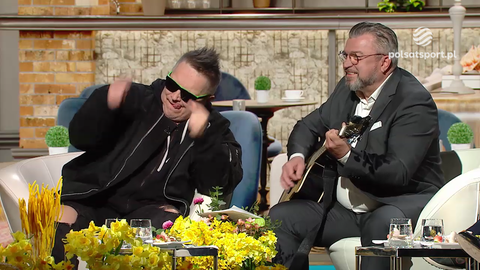Muniek Staszczyk w okularach przeciwsłonecznych i czarnej bluzie gestykuluje, siedząc obok Romana Koseckiego, kt&oacute;ry gra na gitarze akustycznej w studiu telewizyjnym.