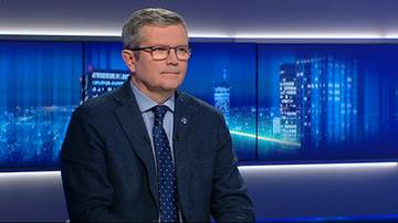 Mężczyzna w garniturze i krawacie, w okularach, siedzi, z poważną miną.