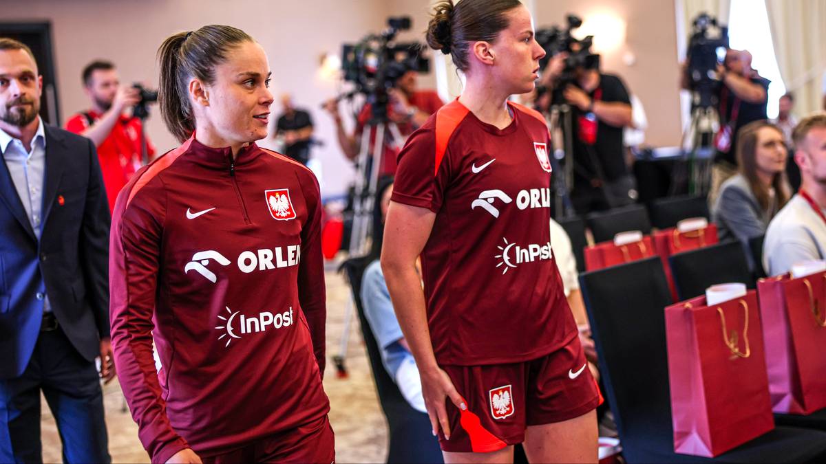 Dwie piłkarki reprezentacji Polski w bordowych strojach, jedna z nich patrzy w lewo, druga w prawo. W tle kamery i ludzie.