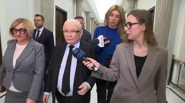 Jarosław Kaczyński w garniturze i krawacie, otoczony przez inne osoby, odpowiada na pytanie dziennikarki z mikrofonem w ręku. W tle widoczny jest jasny korytarz.