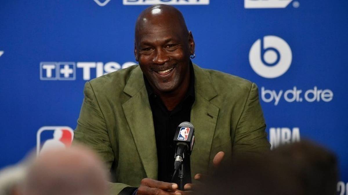 Michael Jordan siedzący za mównicą, uśmiechnięty, przed niebieskim tłem z logo marek.