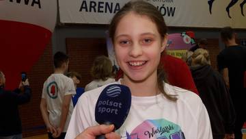 Uśmiechnięta młoda dziewczyna, Hanna Banasik, w białej koszulce z logo "Wieliczka" trzyma mikrofon Polsat Sport. W tle widać ludzi na trybunach i halę sportową.