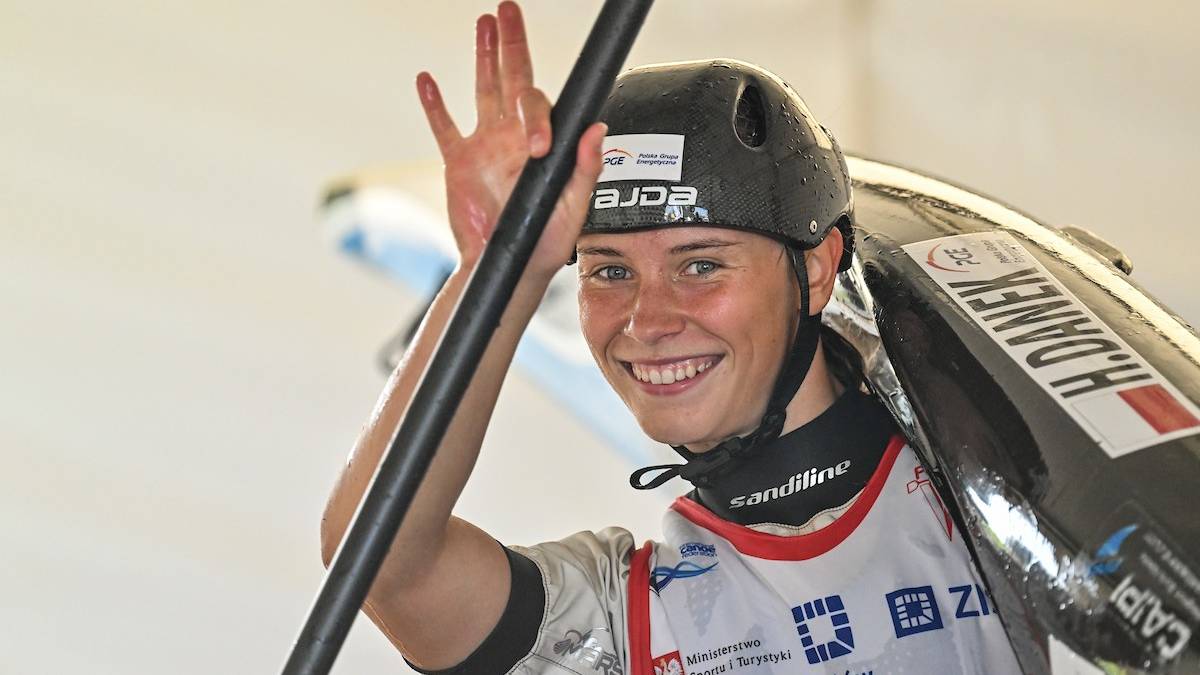 Uśmiechnięta młoda kobieta w kasku kajakarskim i kamizelce sportowej, trzymająca wiosło i unosząca dłoń w geście pozdrowienia.