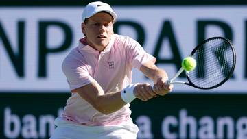 Mężczyzna w białej czapce i r&oacute;żowej koszulce gra w tenisa, uderzając piłkę rakietą.