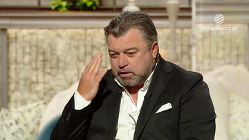 Mężczyzna w czarnym garniturze i białej koszuli siedzi i rozmawia, dotykając dłonią skroni.