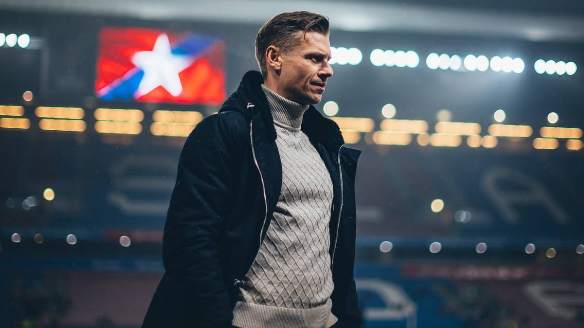 Mężczyzna w zimowej kurtce i golfie stoi na boisku piłkarskim, z tyłu widoczne są światła stadionu i telebim