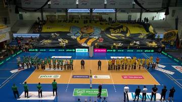 Drużyny siatkarskie stoją po przeciwnych stronach boiska przed rozpoczęciem meczu, z widownią w tle i transparentem.