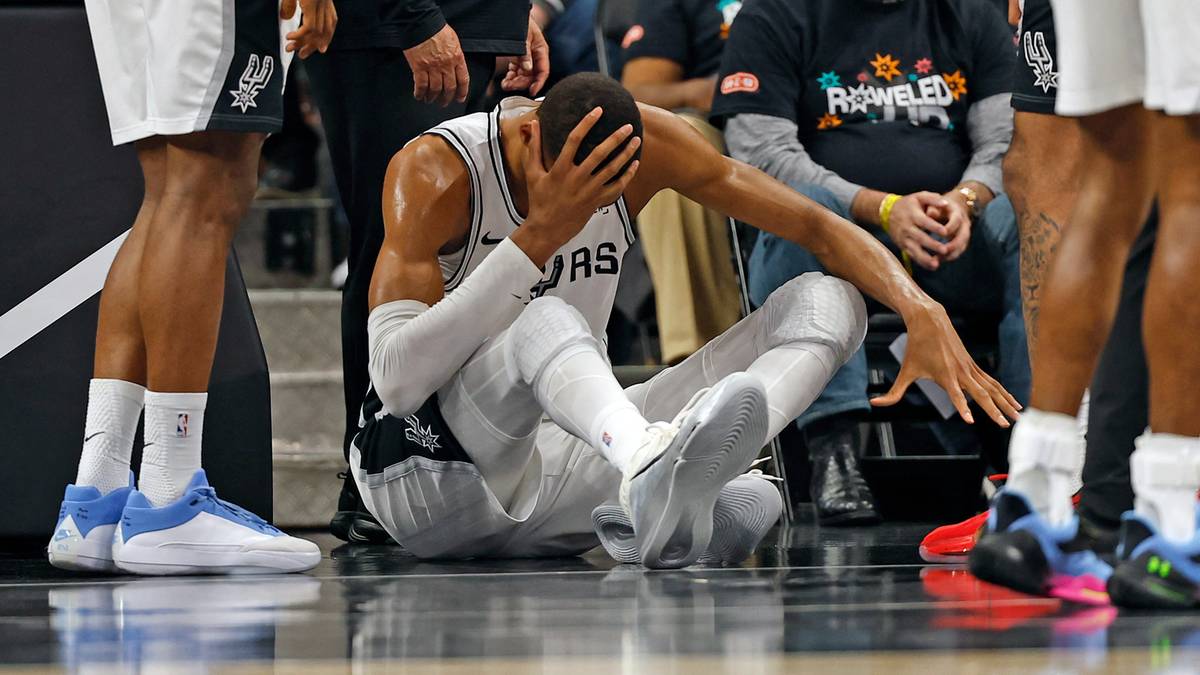 Gracz koszykówki San Antonio Spurs siedzi na parkiecie z ręką na głowie, wyglądając na zranionego.
