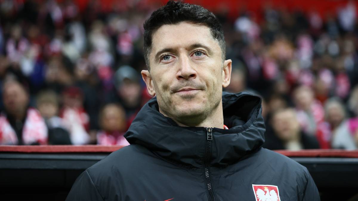 Portret Roberta Lewandowskiego w czarnej kurtce z herbem reprezentacji Polski na tle rozmytych kibiców.