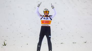 Skoczek narciarski w stroju, z nartami na nogach, unosi ręce w geście triumfu.