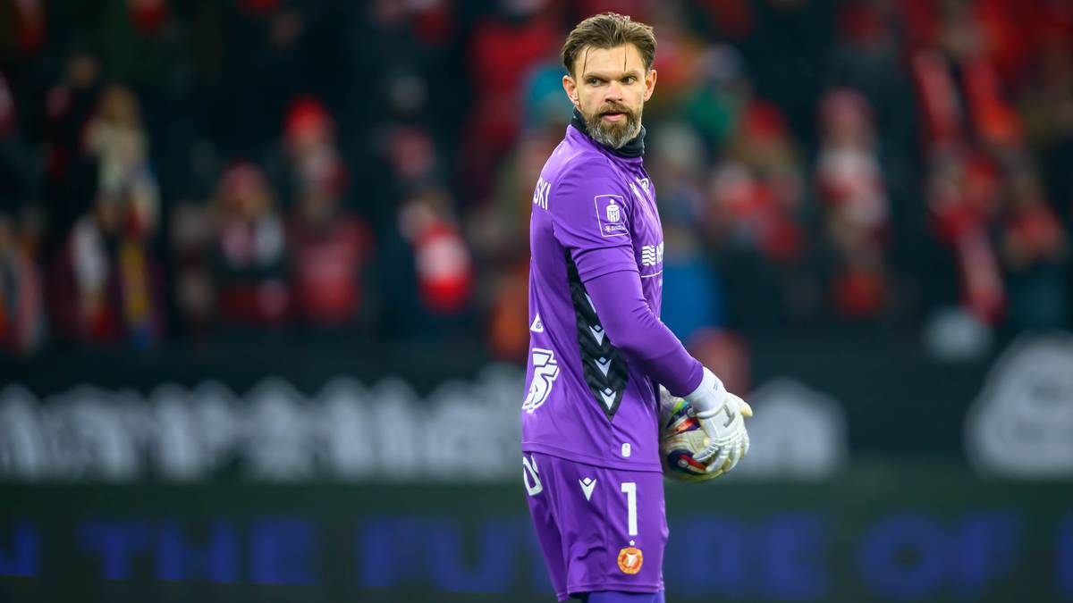 Bramkarz w fioletowym stroju z numerem 1 na koszulce, trzymający piłkę w rękawicach.