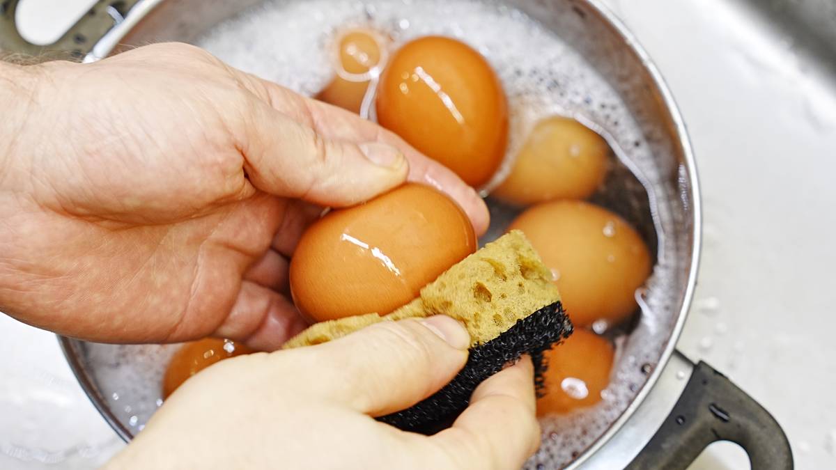 Dłonie trzymające jajka i gąbkę, myjące jajka w garnku z wodą.