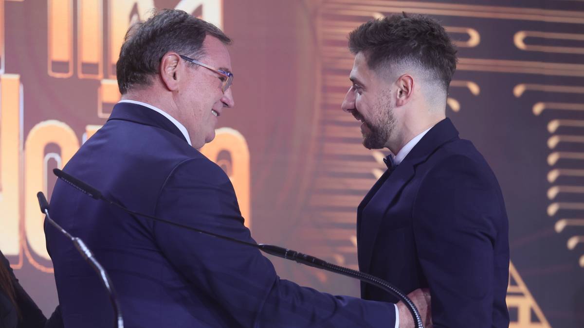 Dw&oacute;ch mężczyzn w eleganckich garniturach stoi przy m&oacute;wnicy, jeden z nich wręcza nagrodę drugiemu. W tle widoczna jest kobieta.