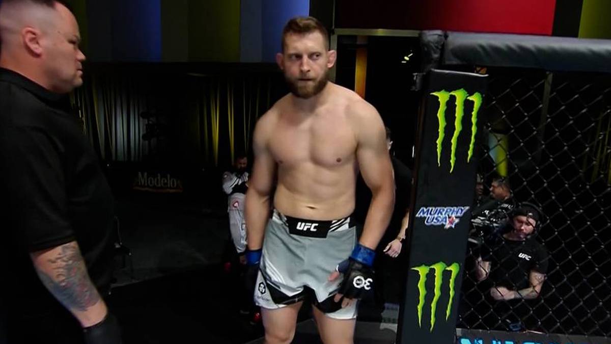 Mężczyzna z zarostem, ubrany w spodenki do MMA i rękawice, stoi przed klatką UFC. Na klatce widnieje logo "Monster Energy". W tle widać ludzi.