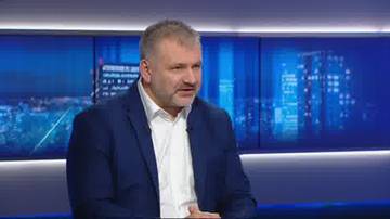 Mężczyzna w białej koszuli i granatowej marynarce m&oacute;wi, siedząc przed ekranem z widokiem na nocne miasto.