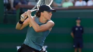 Tenisistka z rakietą tenisową w trakcie zagrania, na tle trybun z widzami.