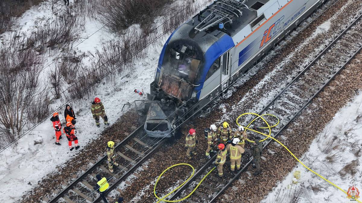 Widok z g&oacute;ry na miejsce wypadku kolejowego, gdzie pociąg zderzył się z samochodem osobowym, kt&oacute;ry zapalił się. Na torach widoczni są strażacy.