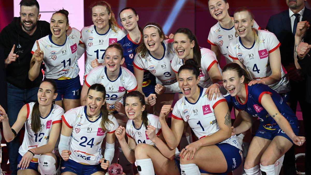 Grupa uśmiechniętych siatkarek w strojach sportowych świętująca zwycięstwo na parkiecie.