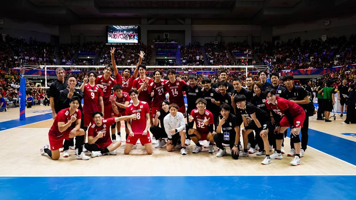 Drużyna siatkarzy w czerwonych strojach pozuje do zdjęcia na tle kibiców na trybunach.