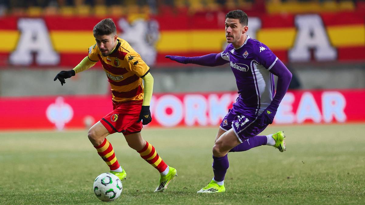 Dw&oacute;ch piłkarzy rywalizujących o piłkę na boisku, jeden w ż&oacute;łto-czerwonym stroju, drugi w fioletowym