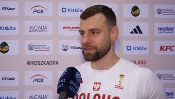 Portret Kamila Łączyńskiego, siatkarza w białej koszulce reprezentacji Polski, z mikrofonem przed ustami.