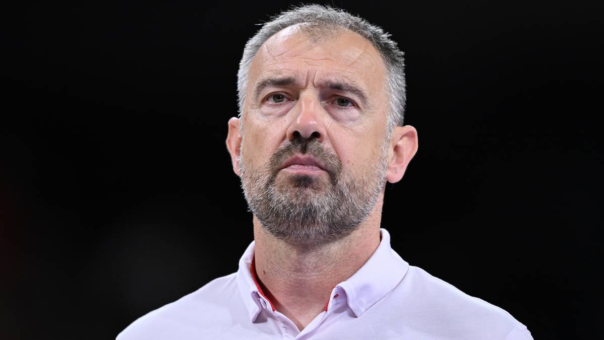 Mężczyzna z siwą brodą i krótkimi włosami, ubrany w jasnoróżową koszulkę polo, patrzy prosto przed siebie z poważną miną.