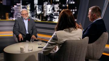 Profesor Szymon Malinowski siedzi przy stole w studiu telewizyjnym, rozmawiając z kobietą i mężczyzną. Na stole stoją szklanki i filiżanki.