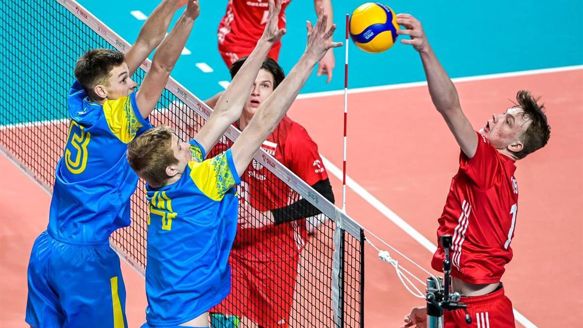 Siatkarz w czerwonej koszulce atakuje piłkę nad siatką, blokowany przez dw&oacute;ch zawodnik&oacute;w w niebiesko-ż&oacute;łtych strojach.