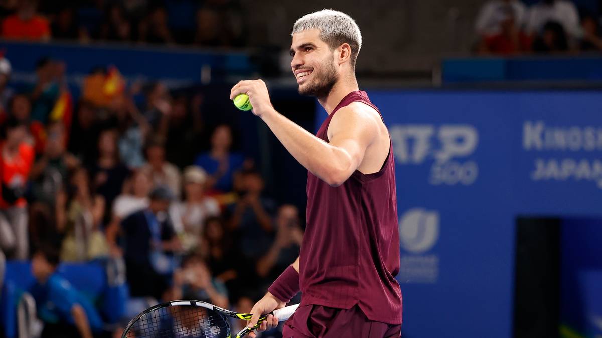 Carlos Alcaraz, uśmiechnięty tenisista z blond włosami, w burgundowej koszulce sportowej, trzymający żółtą piłeczkę tenisową w prawej dłoni, na tle trybun z widzami.
