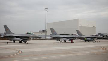 Trzy szare myśliwce F-16 stojące na lotnisku, z białym budynkiem w tle i pochmurnym niebem nad nimi.
