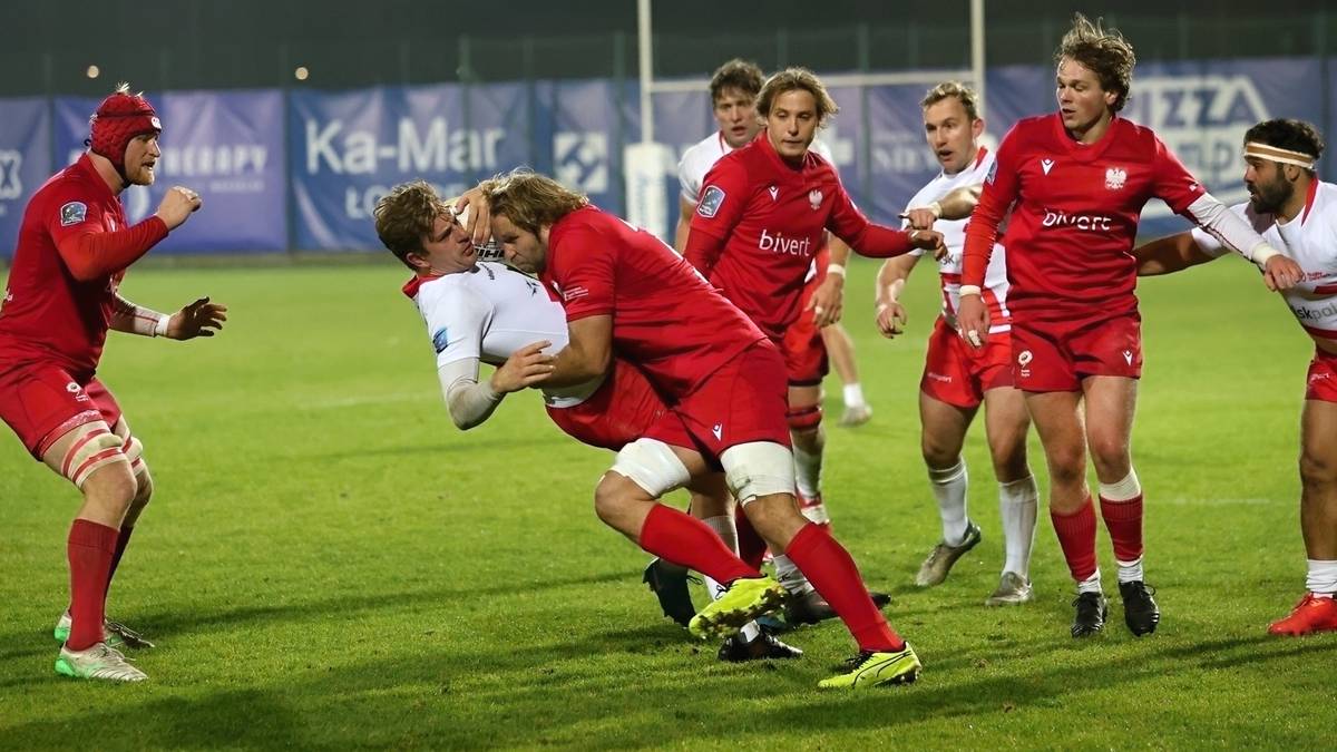 Zawodnicy drużyny rugby walczą o piłkę na boisku, jeden z graczy w czerwonym stroju pr&oacute;buje powalić przeciwnika w białej koszulce.