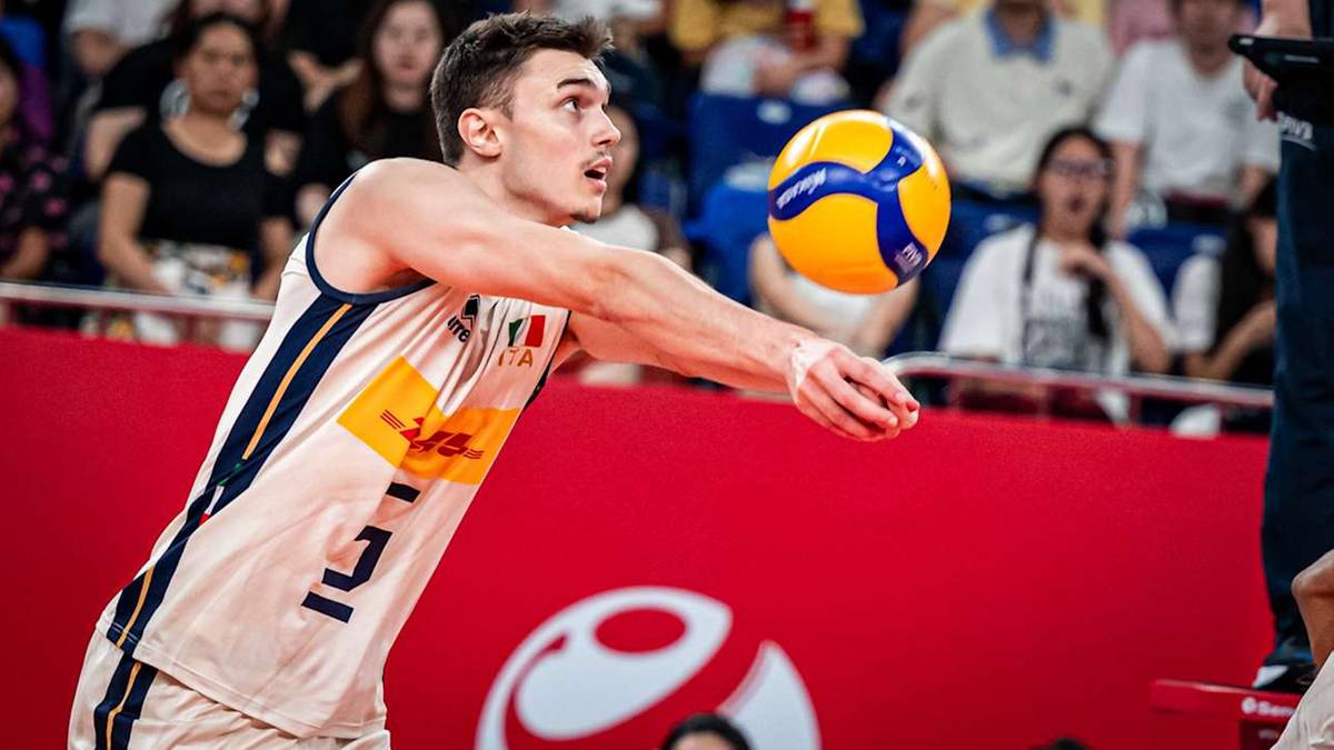 Siatkarz w białej koszulce z elementami graficznymi i numerem 10, wykonuje przyjęcie piłki, blokując ją przed sobą obiema rękami. Piłka jest żółto-niebieska.