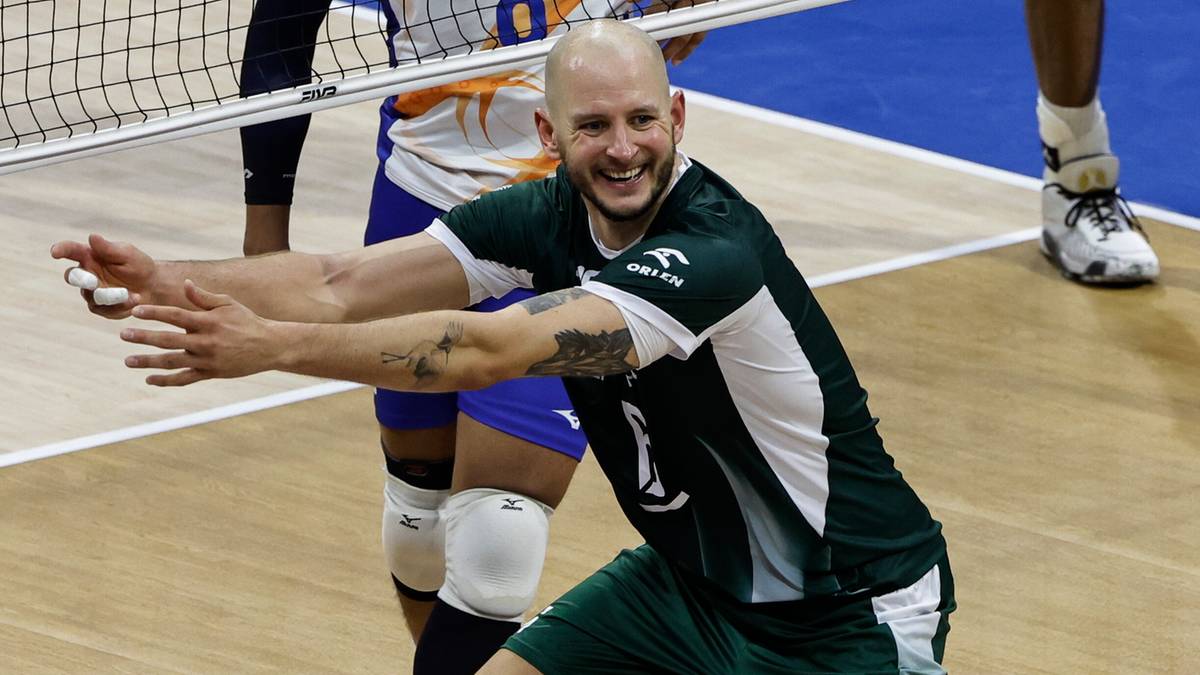 Siatkarz Bartosz Kurek z szerokim uśmiechem, patrzący w lewo, w zielono-białej koszulce sportowej, ze zgiętymi rękami, podczas meczu siatk&oacute;wki.