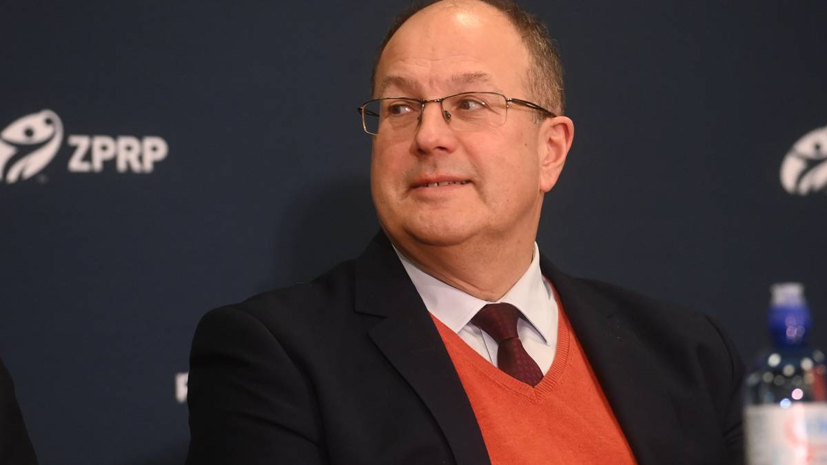 Portret mężczyzny w okularach, w marynarce, białej koszuli i pomarańczowym swetrze, z ciemnym krawatem.