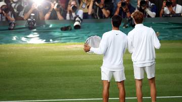 Dwóch tenisistów w białych strojach stoi na zielonym, trawiastym korcie tenisowym, odwróconych plecami. Za nimi widać zgromadzonych ludzi z aparatami fotograficznymi.