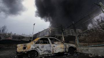 Spalony samoch&oacute;d na tle zniszczonego budynku i kłębu czarnego dymu.