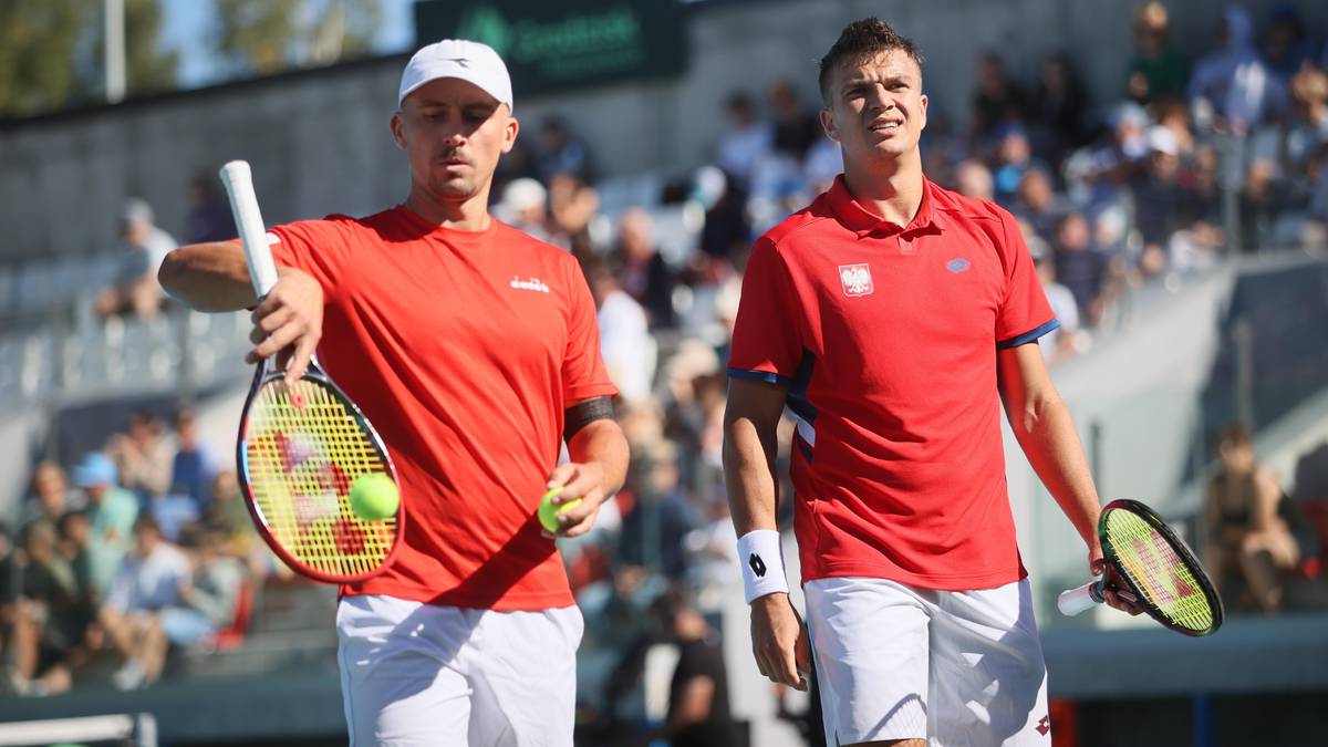 Dwóch tenisistów w czerwonych koszulkach i białych spodenkach stoi obok siebie na korcie tenisowym. Jeden z nich, w białej czapce, trzyma rakietę i piłkę tenisową. Drugi gracz, z wyrazem determinacji na twarzy, również trzyma rakietę. W tle widoczna jest trybuna z widzami.