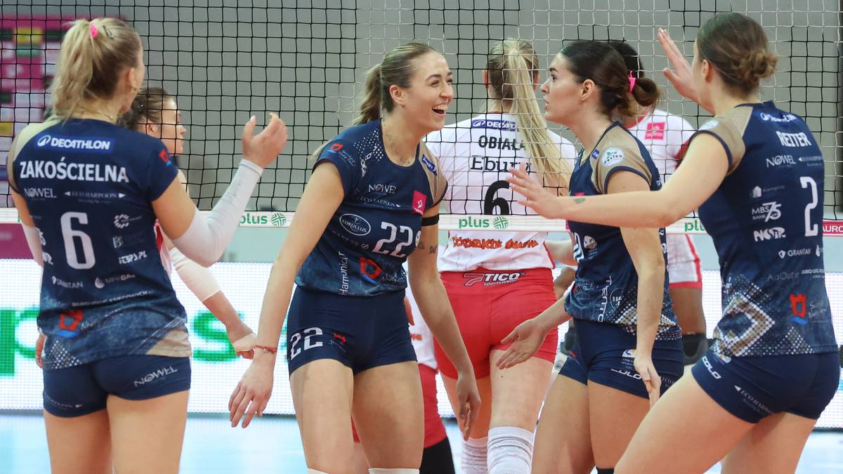 Fragment meczu siatk&oacute;wki, gdzie zawodniczki w ciemnogranatowych strojach rywalizują na boisku, dwie z nich rozmawiają ze sobą.
