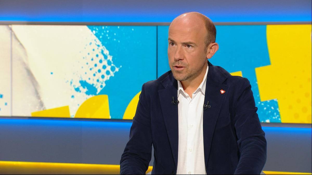 Mężczyzna w granatowym garniturze i białej koszuli, patrzący przed siebie. W tle widoczne są abstrakcyjne, kolorowe panele w odcieniach żółci, niebieskiego i bieli.