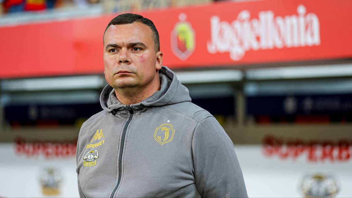 Mężczyzna w szarej bluzie z kapturem z logo Jagiellonii Białystok, stojący na tle stadionu.