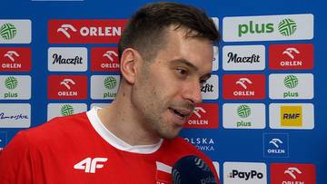 Marcin Komenda w czerwonej koszulce reprezentacji Polski udziela wywiadu na tle ścianki sponsorów.