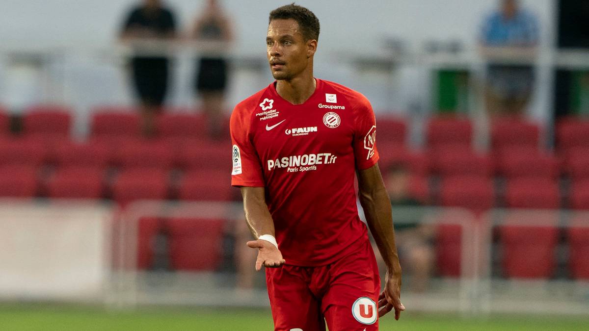 Portret piłkarza w czerwonej koszulce meczowej, stojącego na boisku z gestem otwartej dłoni. W tle widać czerwone trybuny stadionu.