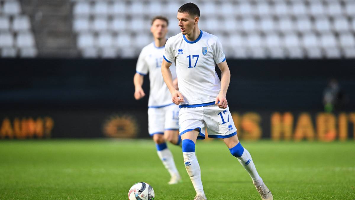 Piłkarz w białej koszulce z numerem 17 i niebieskimi detalami biegnie z piłką na zielonej murawie stadionu.